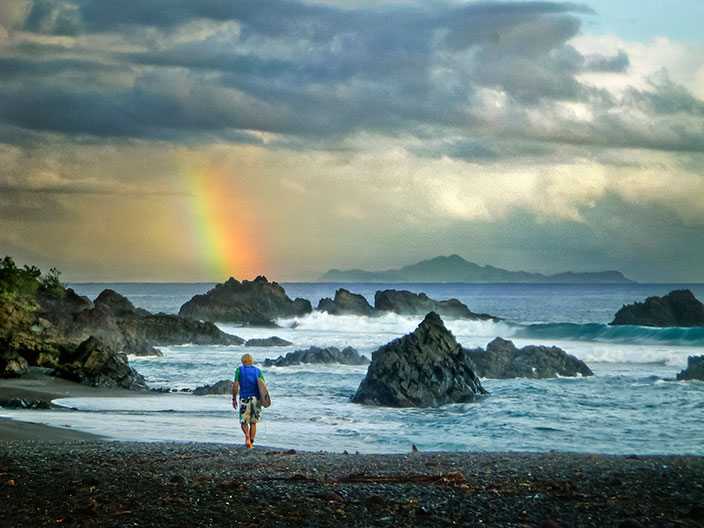 Cover-More New Zealand Facebook photo competition winner: Surfer walks towards the beach as a rainbow appears above him